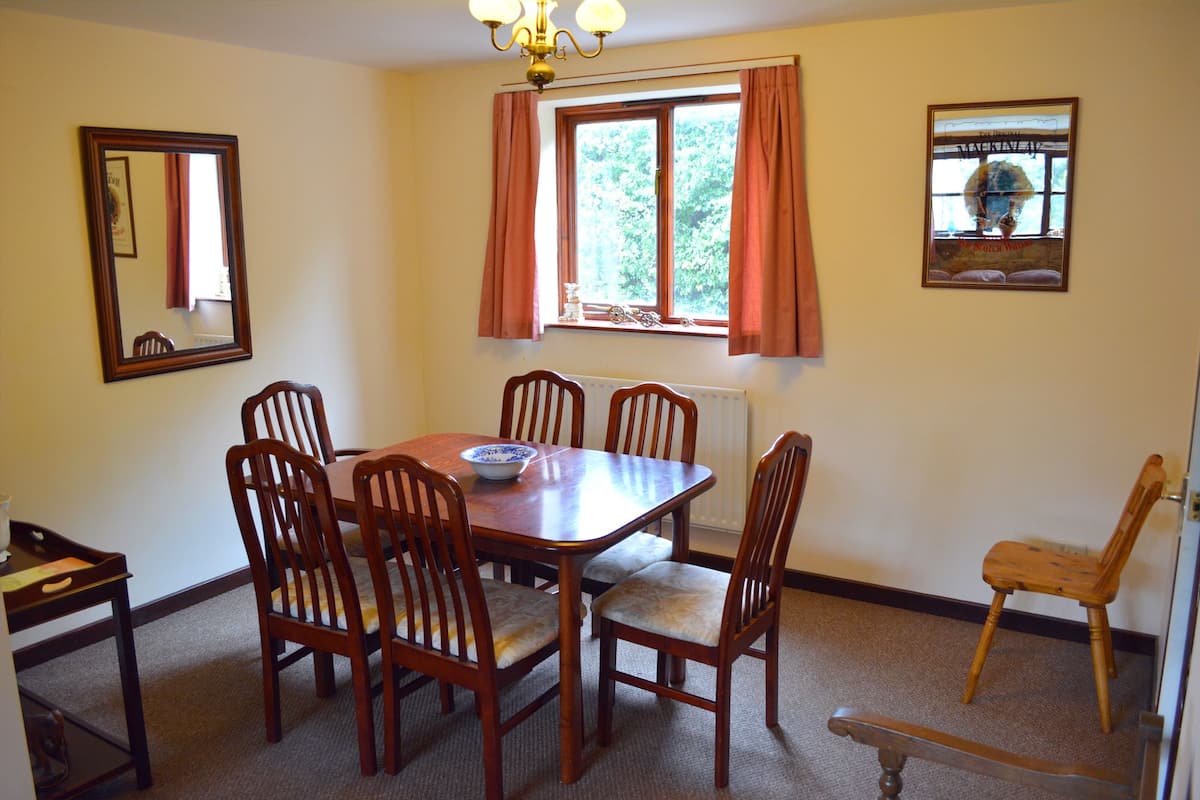A dining room with a wooden table and chairs, a radiator, a window with curtains, and a framed picture on the wall.