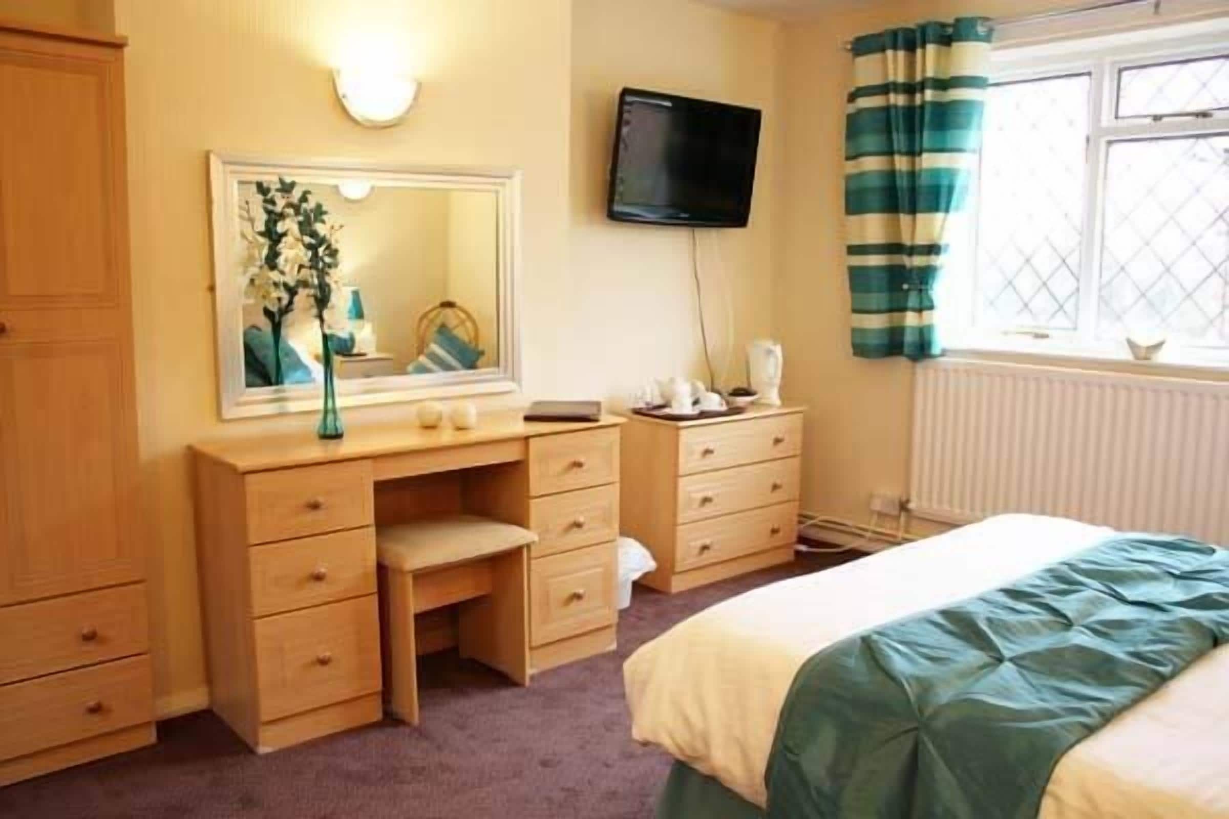 A bedroom with a bed, a desk, a mirror, a television, and a window with curtains.