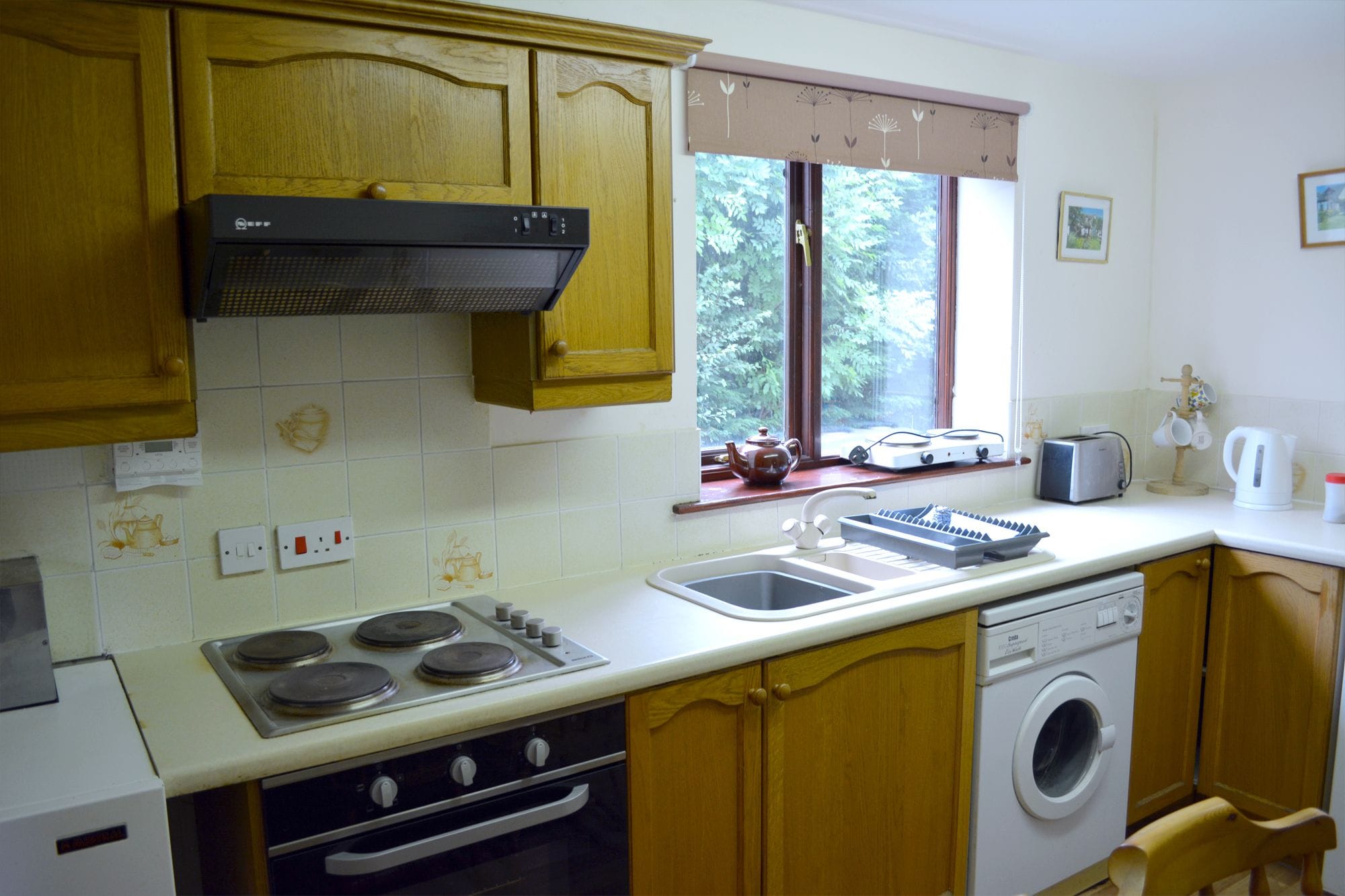 A kitchen with wooden cabinets, a stove, a sink, a window with a view of greenery, and a washing machine.