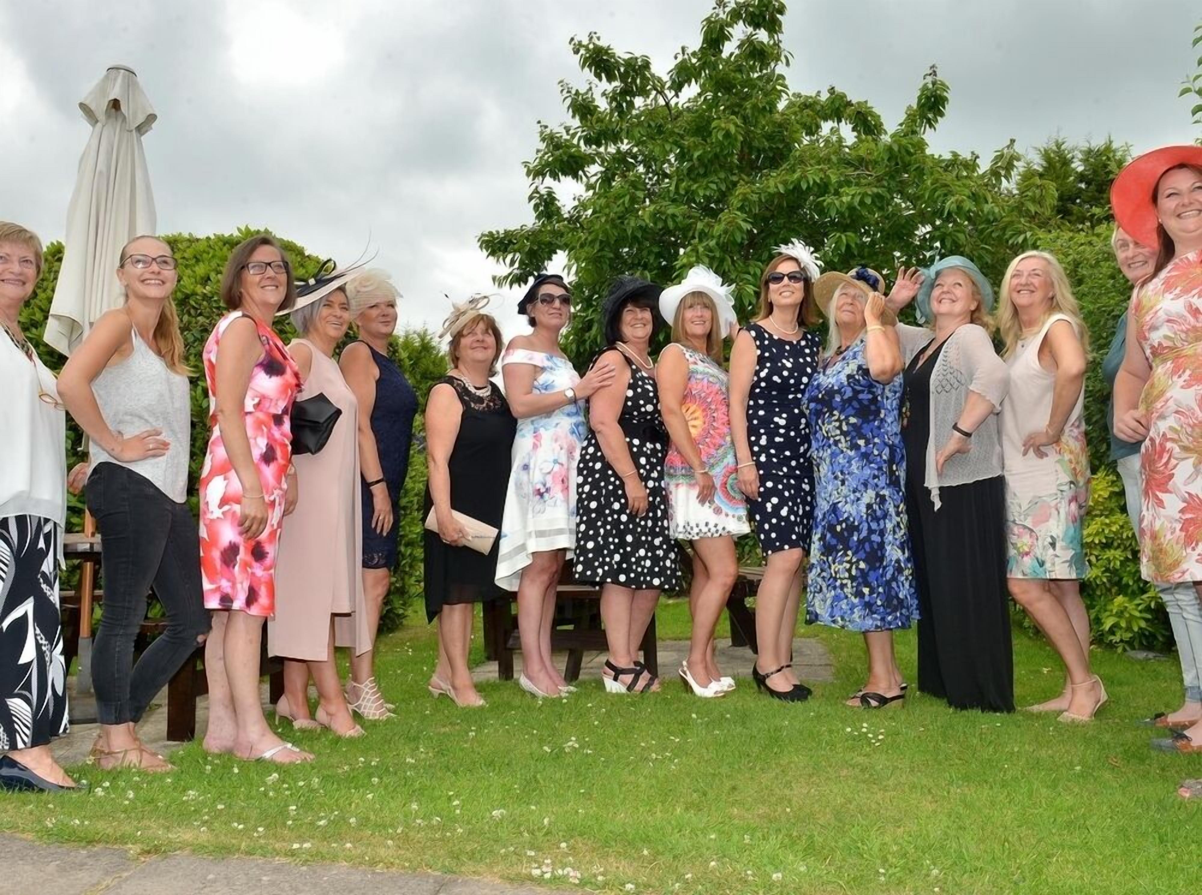 A group of people standing outdoors, possibly at a social event, with a grassy area and trees in the background.