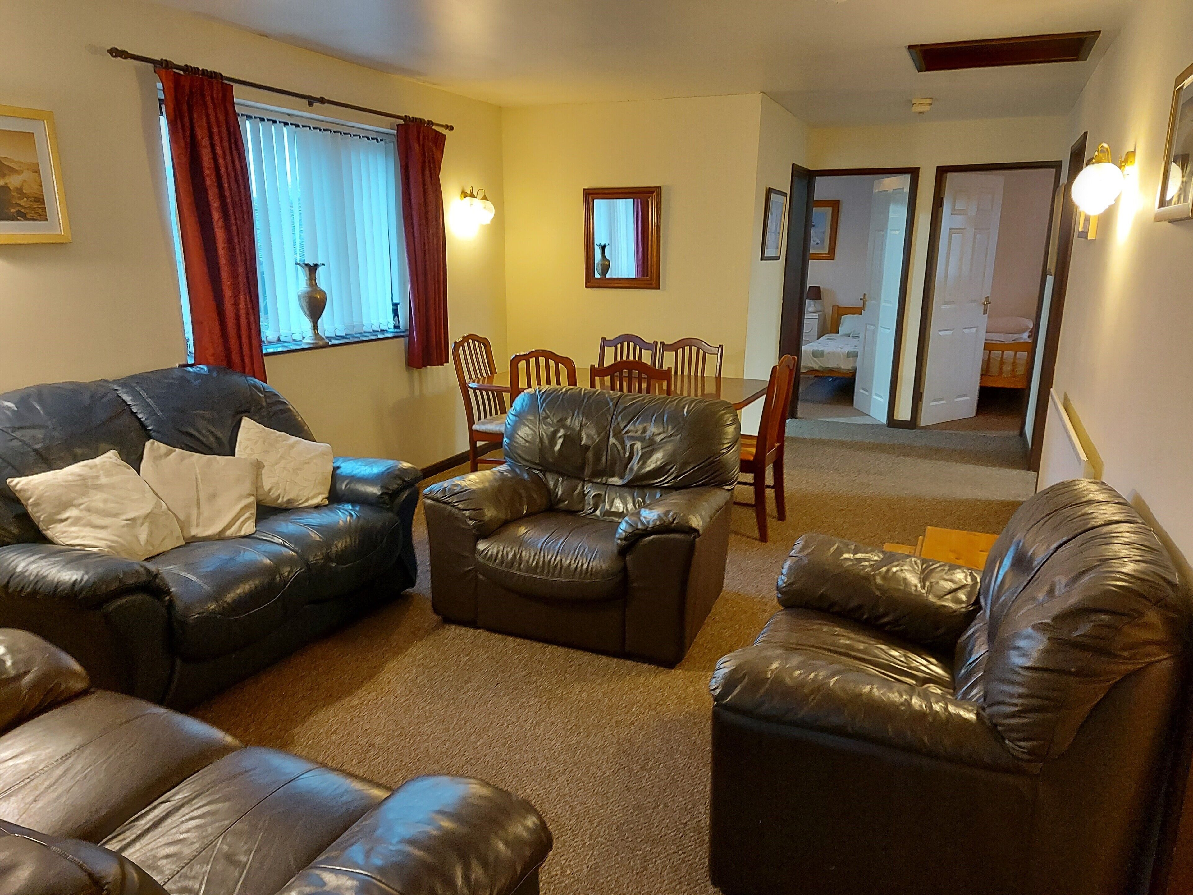 A living room with leather sofas, a dining table with chairs, and a hallway leading to a bedroom.
