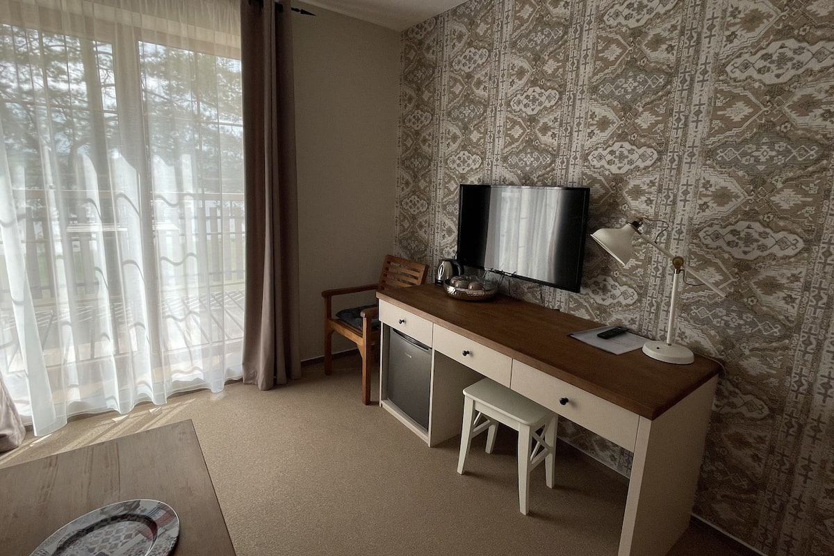 Habitación de hotel con un escritorio de madera, televisor, un mueble pequeño, una silla, un banco y un papel tapiz estampado.