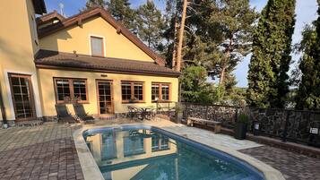 A swimming pool with a stone border, surrounded by a paved patio and a house with a tiled roof and windows.