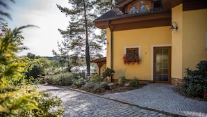 A cobblestone driveway leading to a yellow building with a wooden door and window, surrounded by greenery and trees. - Resort-Restaurant Štilec (Kamenný Újezd)