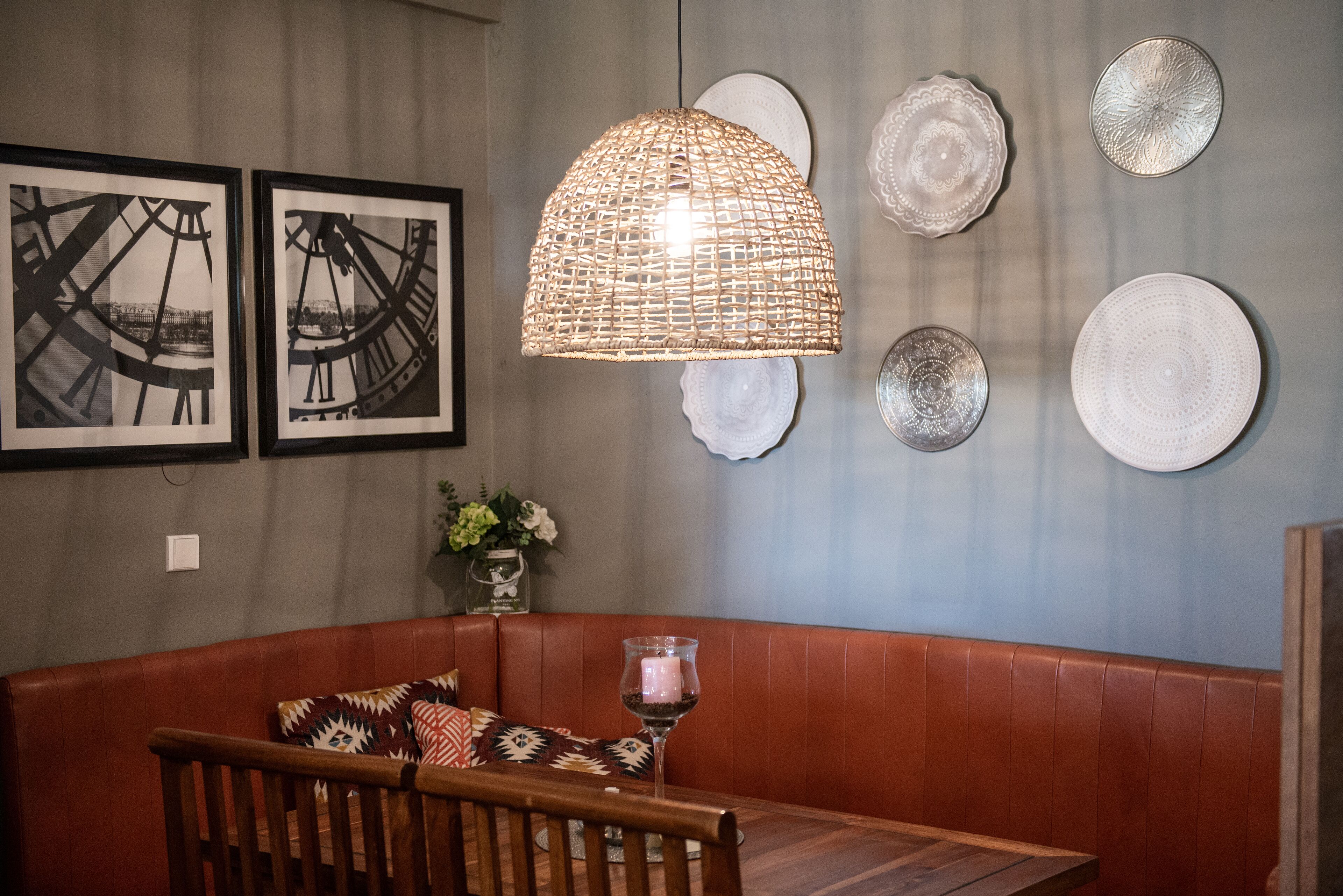 A dining area with a wooden table, a cushioned bench, a woven pendant light, and framed artwork on the wall.