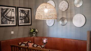 A dining area with a wooden table, a cushioned bench, a woven pendant light, and framed artwork on the wall.
