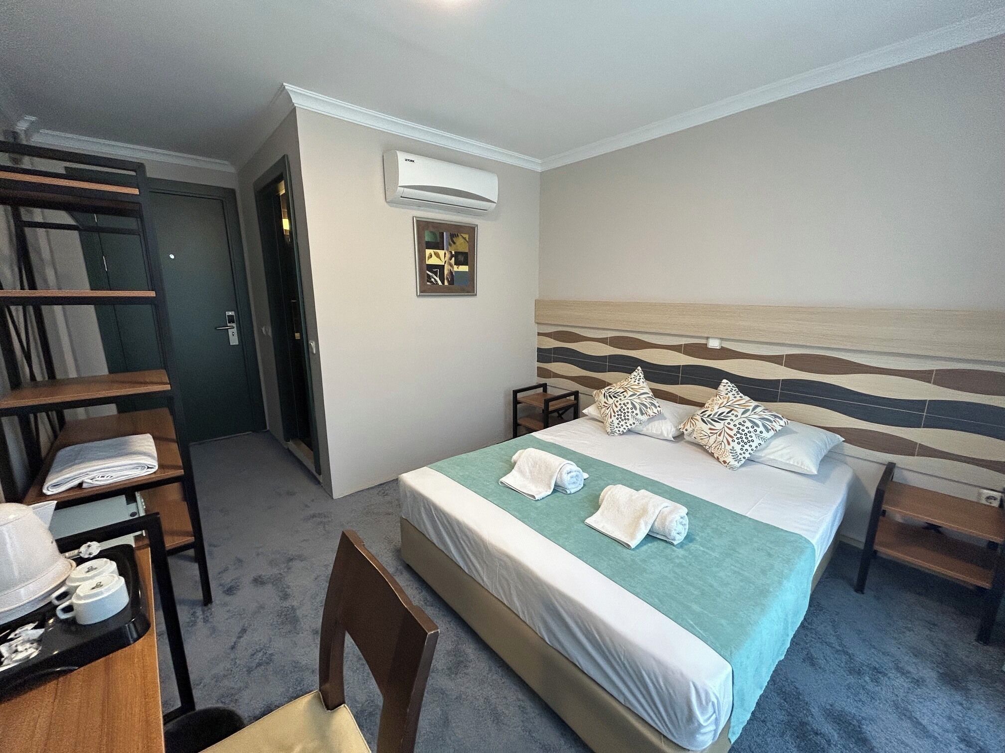 A hotel room with a bed, a chair, a desk with a tray of items, an air conditioning unit, and a door.