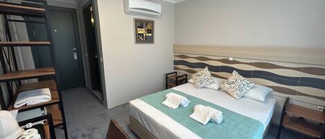 A hotel room with a bed, a chair, a desk with a tray of items, an air conditioning unit, and a door.