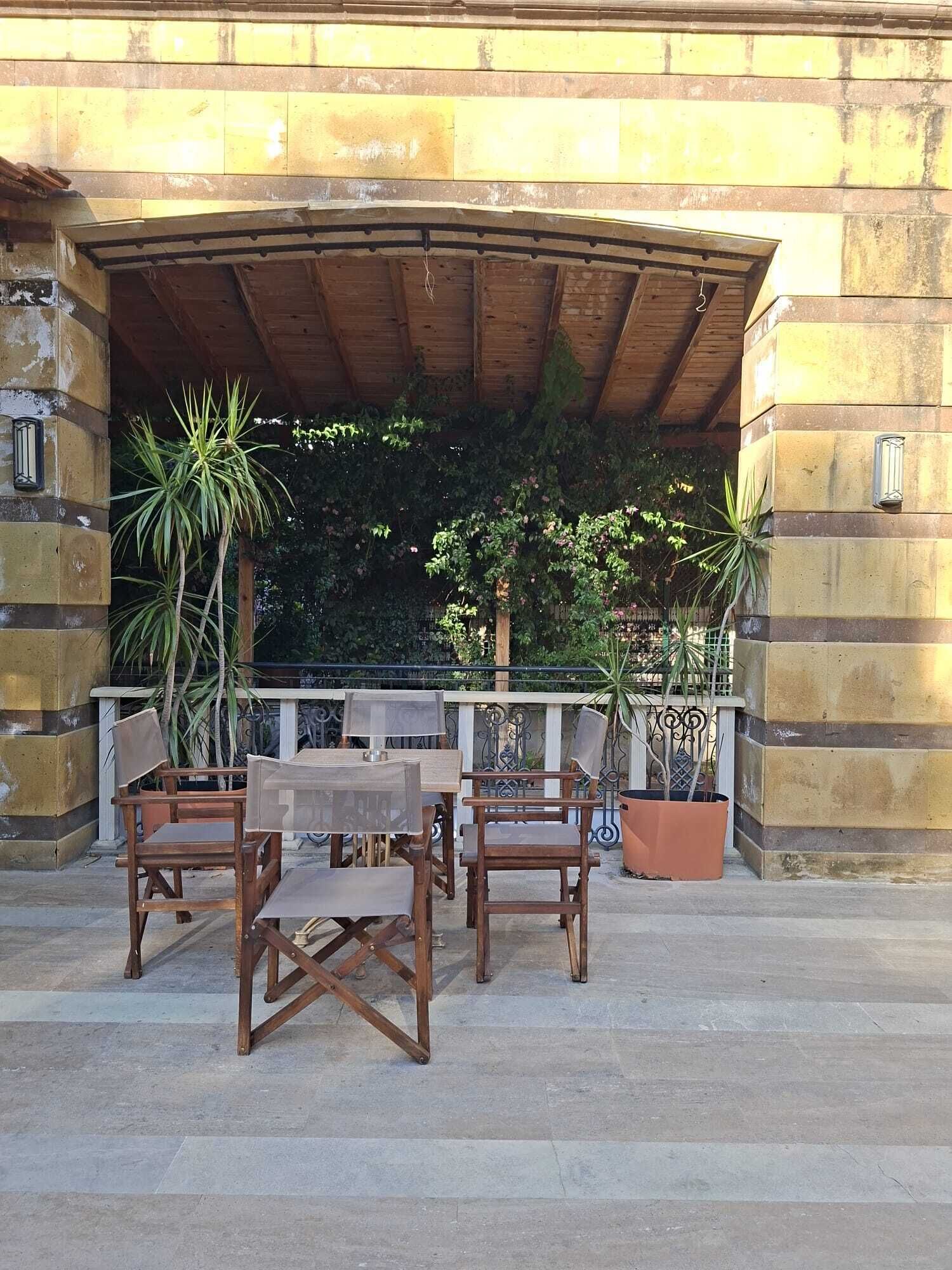 An outdoor patio with a wooden table and chairs, surrounded by potted plants and a stone wall.