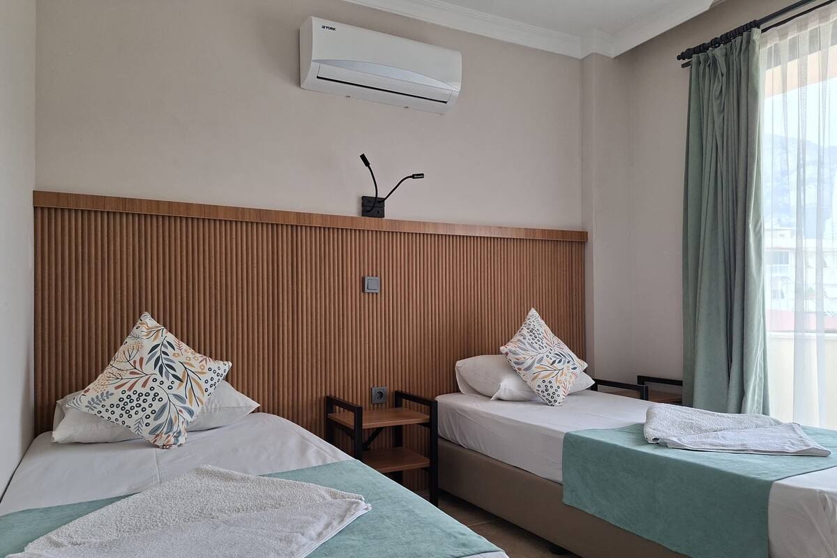 Habitación de hotel con dos camas, cabecera de madera, unidad de aire acondicionado, una mesita pequeña y una ventana con cortinas.