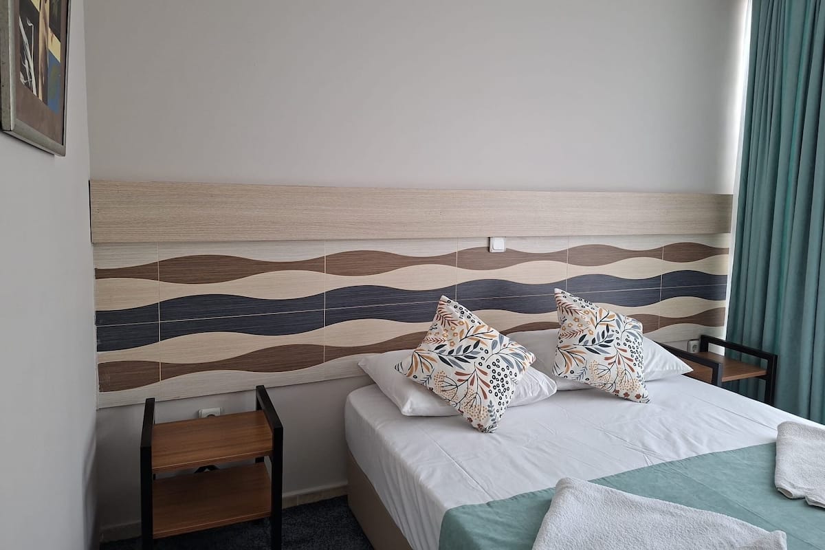 Habitación de hotel con cama, dos almohadas, mesita de noche, una silla y pared con diseño ondulado.