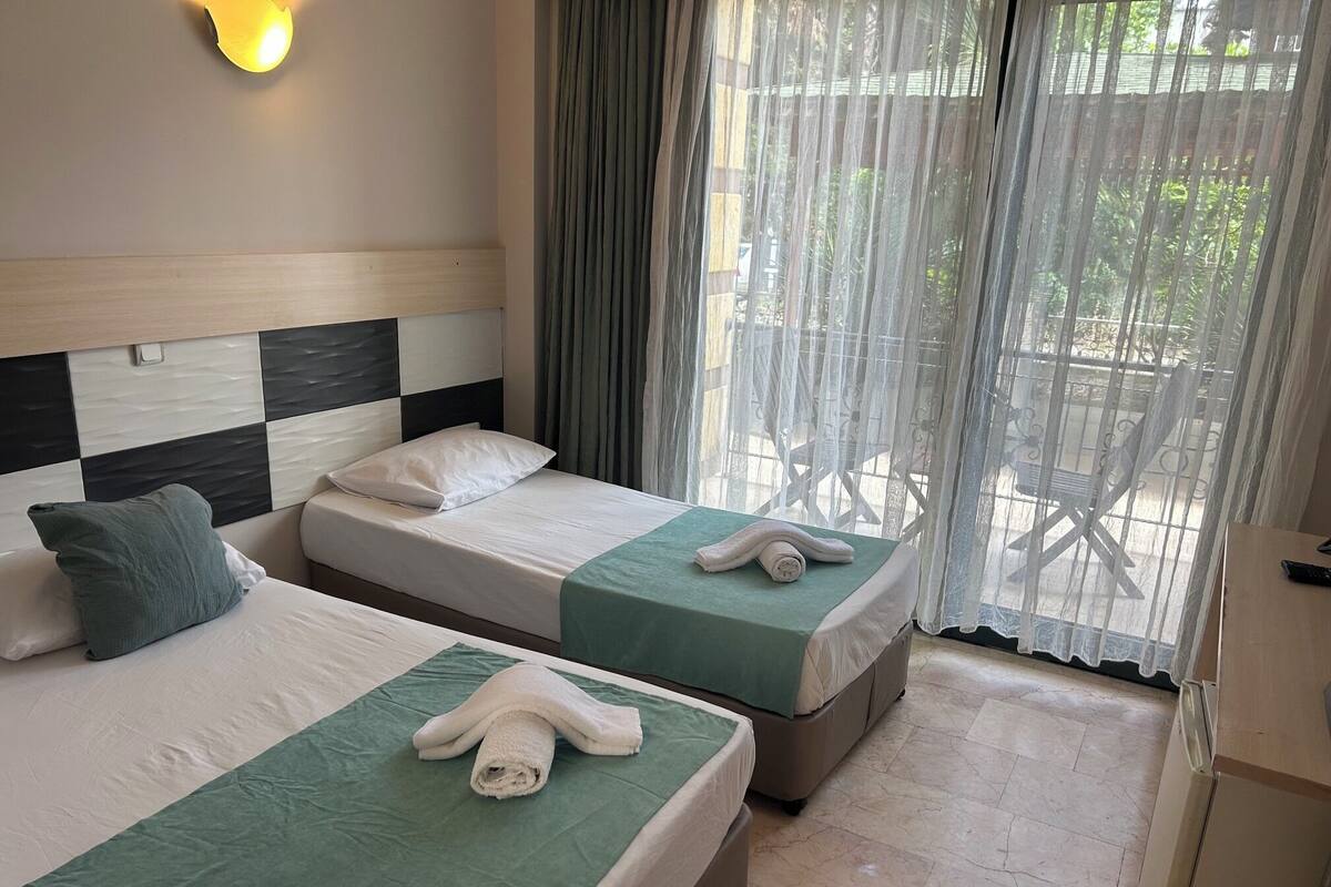 Habitación de hotel con dos camas, una colcha verde, almohadas blancas, una toalla blanca sobre cada cama, un cabecero de madera, una lámpara de pared y vistas al exterior a través de cortinas translúcidas.