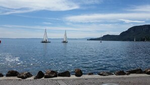 A calm lake with sailboats, a rocky shoreline, and distant mountains. - La Vittoria Boutique Hotel (Garda)