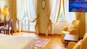 A hotel room with a bed, two yellow armchairs, a small table, a desk with a lamp, and a TV displaying a cityscape. - La Vittoria Boutique Hotel (Garda)