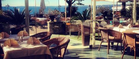 An outdoor dining area with tables and wicker chairs, overlooking a scenic view of the sea and hills.