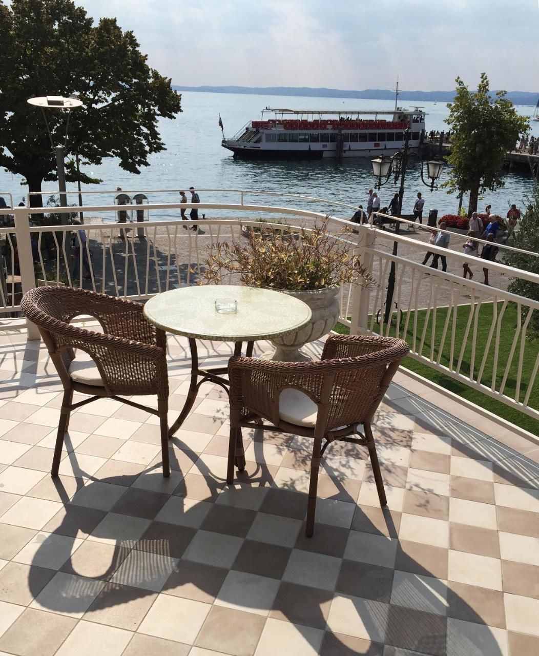 A terrace with wicker chairs and a table, overlooking a body of water with a boat and people in the background.