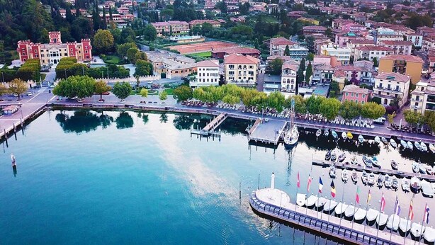 A marina with boats docked, a large building with a tower, and surrounding residential buildings. - La Vittoria Boutique Hotel (Garda)