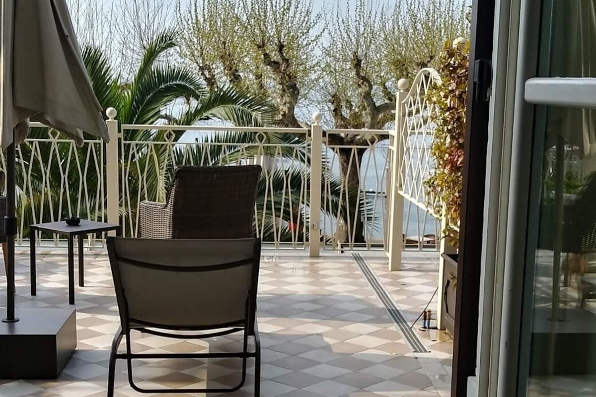 Une terrasse extérieure au sol carrelé, avec une chaise et une table, une porte vitrée et une vue sur des arbres et une clôture.