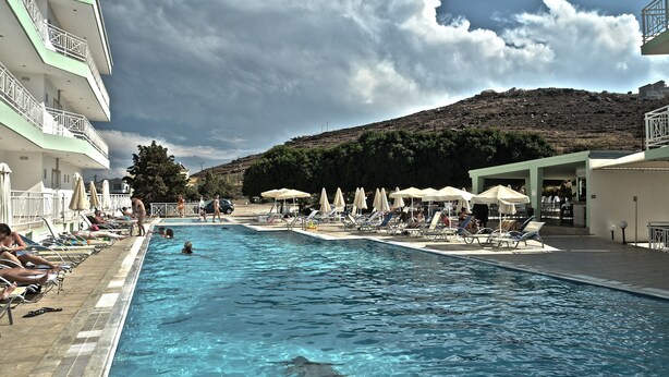 A swimming pool with lounge chairs and umbrellas, surrounded by buildings and a hill. - Chrissy's Paradise (Malevizi)