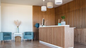 A modern reception area with a wooden panel wall, a reception desk, two blue chairs, a small side table with a vase and a lamp, and three pendant lights. - Chrissy's Paradise (Malevizi)