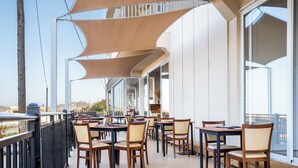 An outdoor dining area with tables and chairs, large windows, and a view of the cityscape. - Chrissy's Paradise (Malevizi)