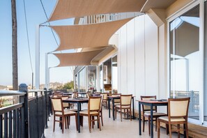 An outdoor dining area with tables and chairs, large windows, and a view of the cityscape. - Chrissy's Paradise (Malevizi)