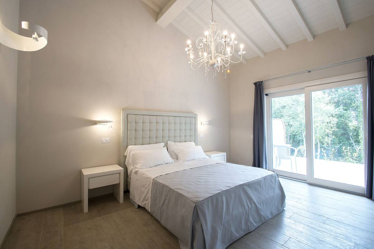 A bedroom with a large bed, a nightstand, a bench, a chandelier, and a large window with curtains.