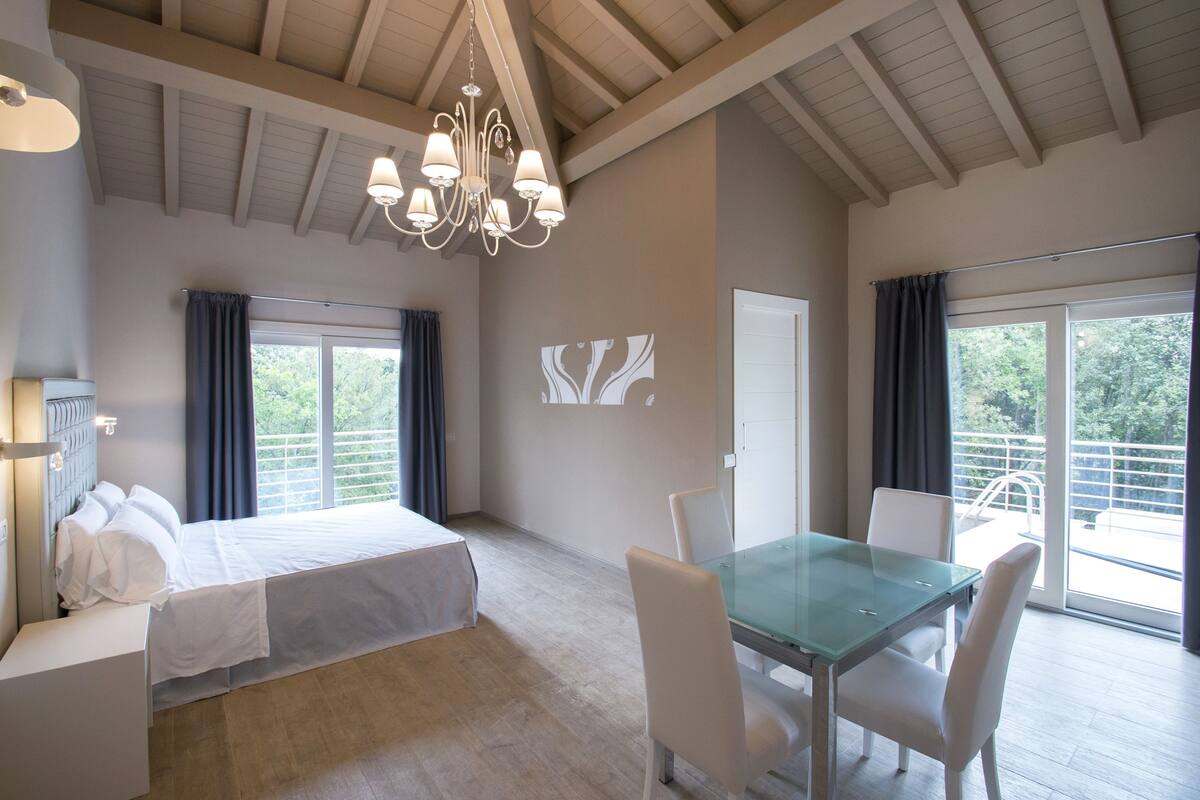 A bedroom with a bed, a glass dining table, and chairs, a chandelier, and a balcony with a railing.