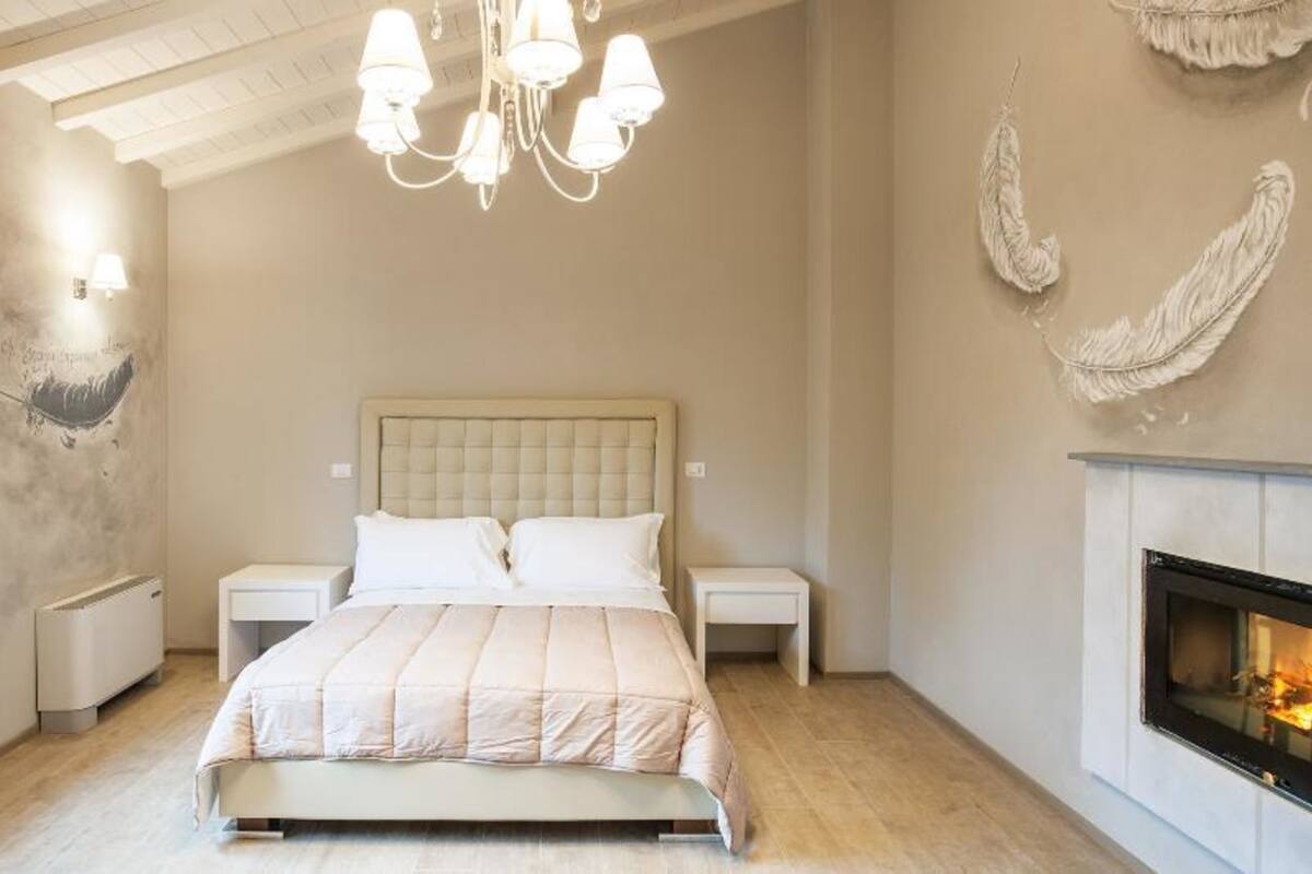 A bedroom with a bed, bedside tables, a chandelier, a fireplace, and wall art.