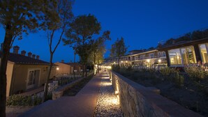 Un chemin bordé d’arbres et délimité par un mur de pierres, qui mène  à un bâtiment doté de grandes fenêtres et d’un éclairage extérieur.
