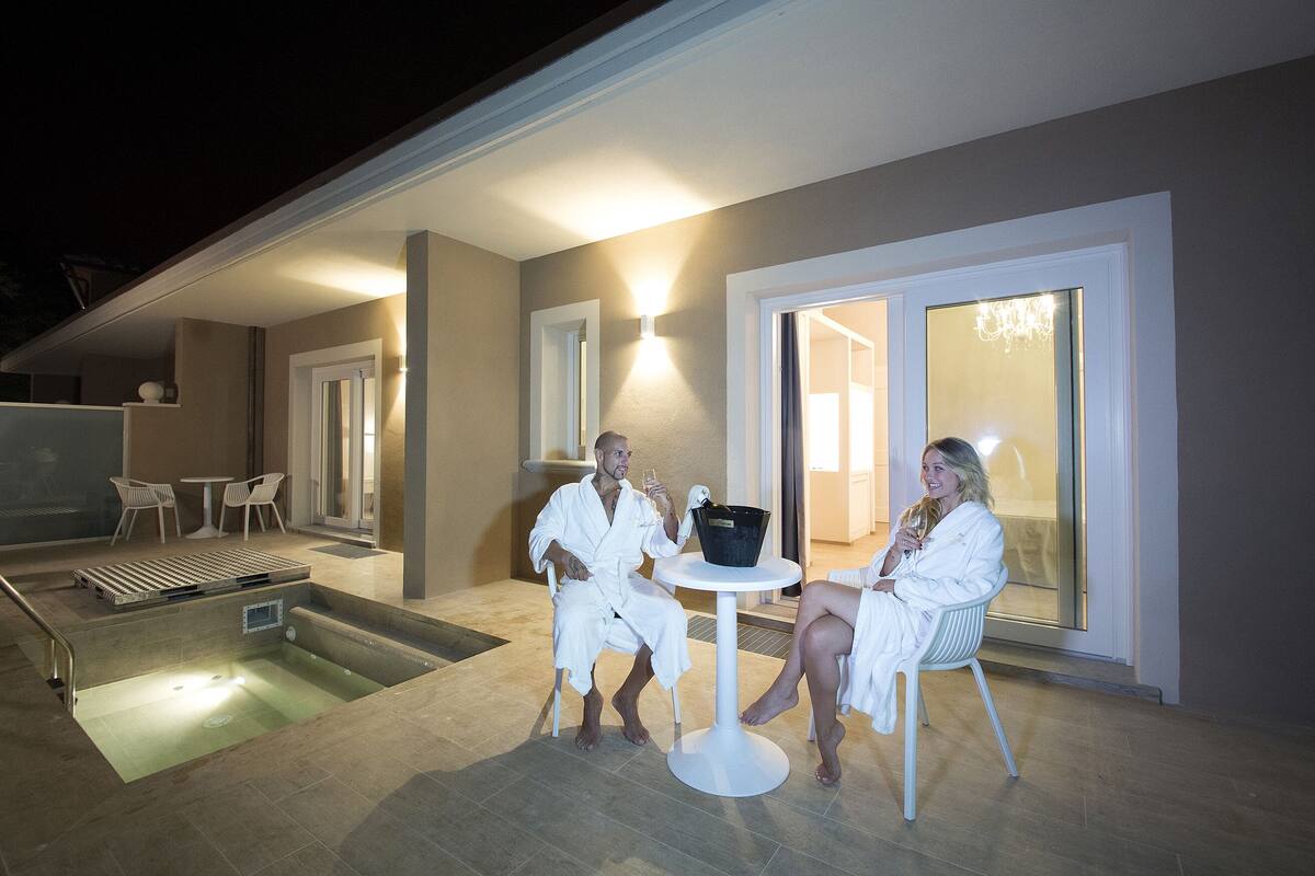 A couple in bathrobes at an outdoor heated pool area with a table and chairs, a small pool, and a covered patio.