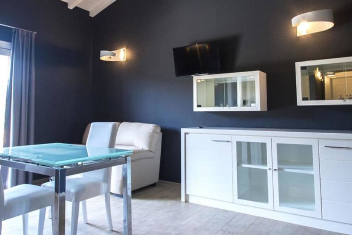 A modern room with a glass table, white chairs, a white sofa, a TV mounted on the wall, and a cabinet with glass doors.
