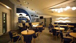 A modern bar area with round tables and blue chairs, a counter with a TV, and a patterned wall. - Grand Laola Spa (Rewal)