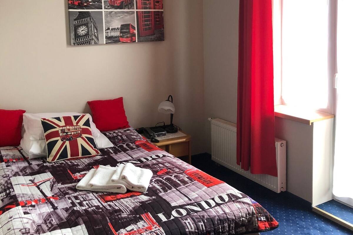 A bedroom with a bed, a nightstand, a lamp, a radiator, and a window with red curtains.
