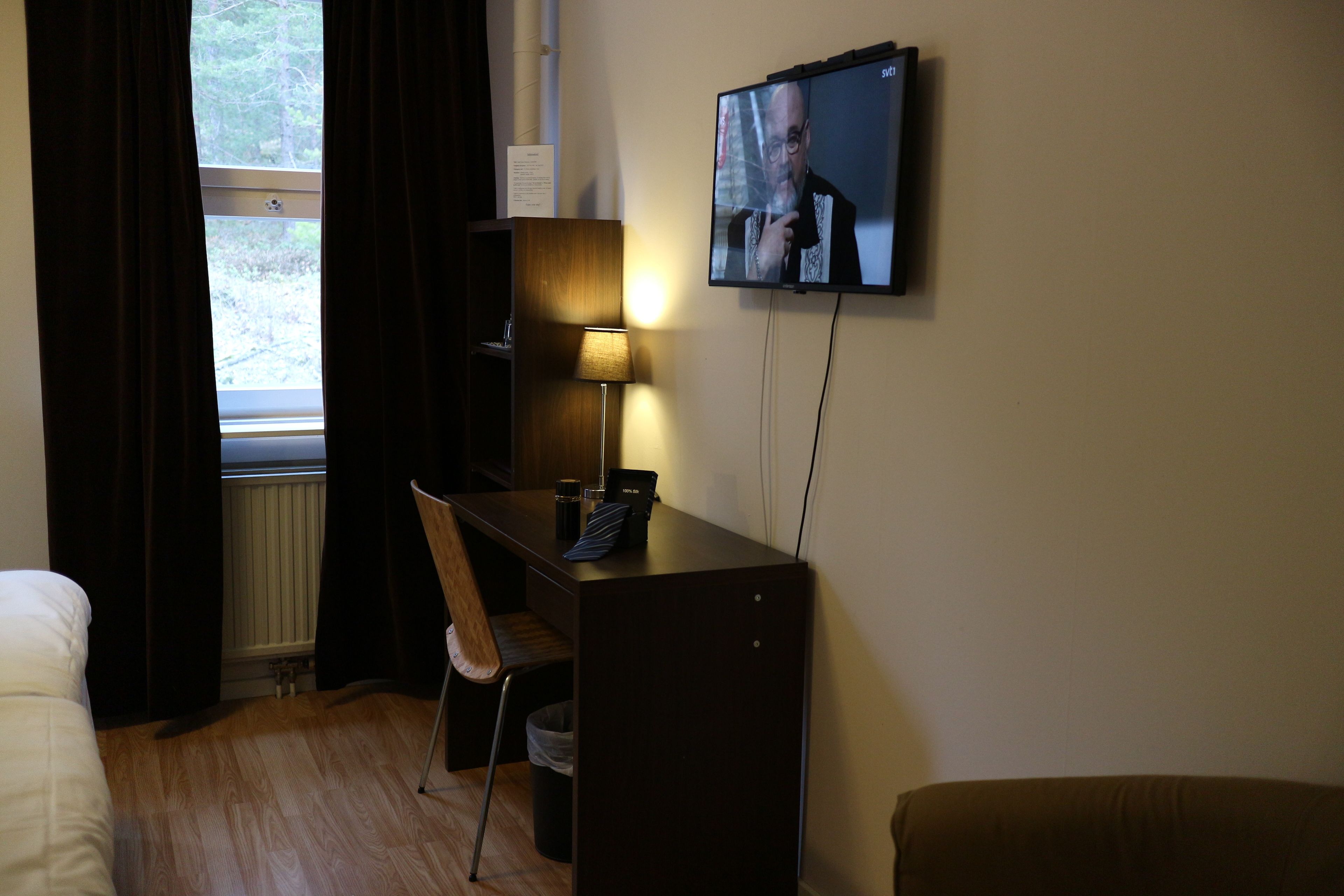 Une chambre d’hôtel comprenant un lit, un bureau avec une chaise, un téléviseur fixé au mur et une fenêtre avec des rideaux.