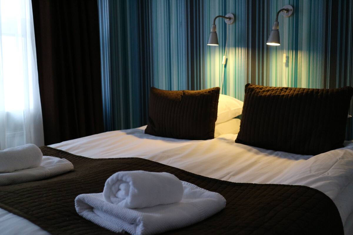A hotel room with a bed, two pillows, a blanket, and a folded towel.