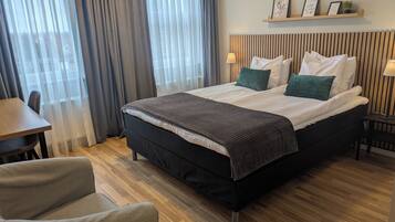 A hotel room with a bed, a desk, a chair, a window with curtains, and a shelf with framed pictures.