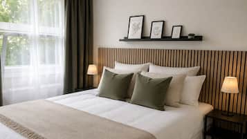 A neatly made bed with a beige blanket and pillows, a bedside table with a lamp, and a wall-mounted shelf with framed botanical prints.