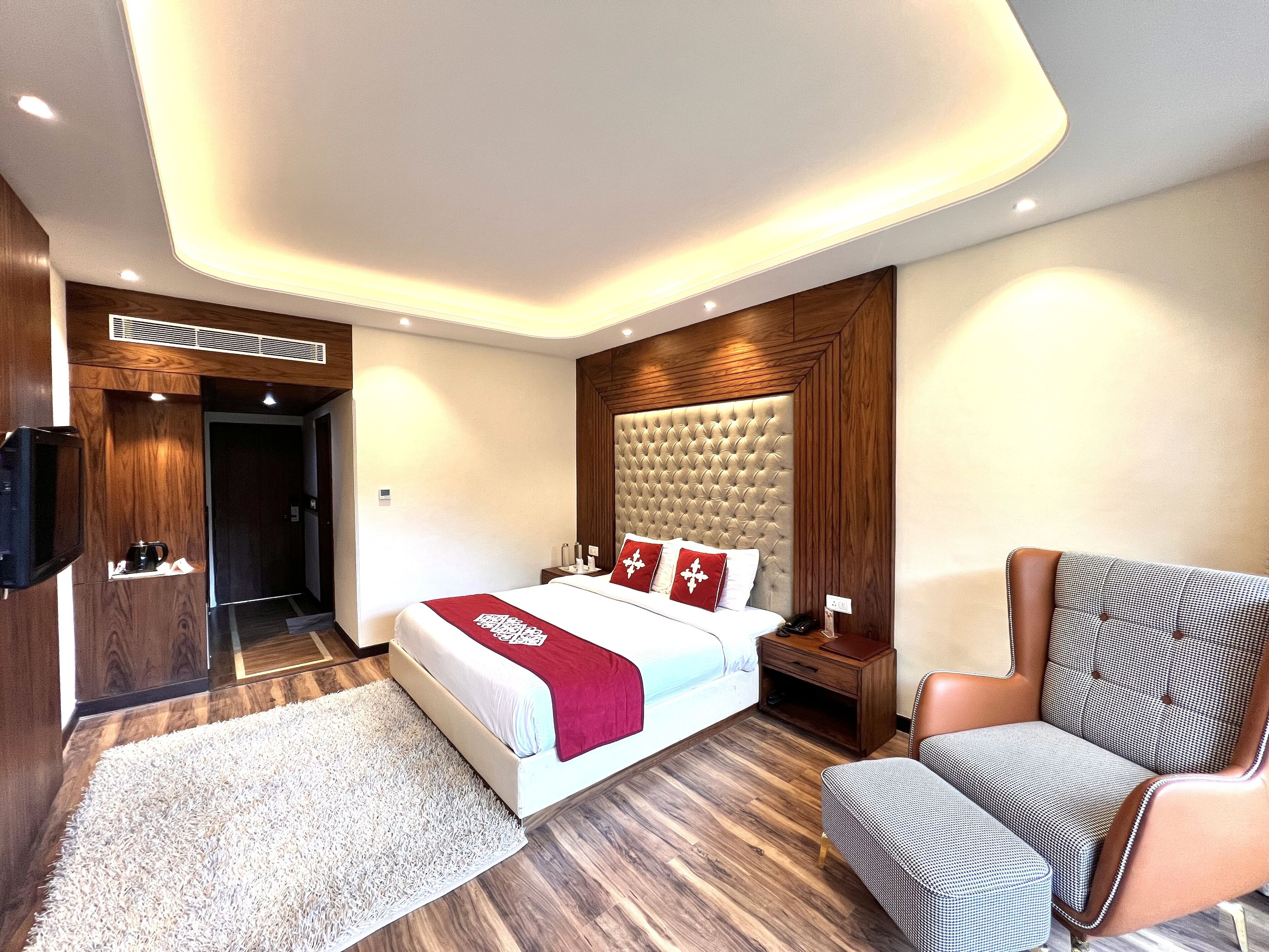 A modern hotel room with a large bed, a wooden headboard, a sitting area with a chair, and a television on the left.