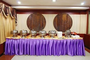 Una mesa de buffet con varios recipientes de comida y una jarra blanca, sobre un fondo de paneles de madera y una cortina con drapeado.
