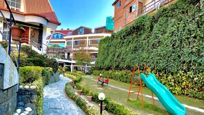 A courtyard with a children's play area, including a slide and swings, surrounded by neatly trimmed hedges and potted plants. - Marigold Sarovar Portico , Shimla. Heated pool & AC Rooms (Resort cottage and SPA) (Shimla)
