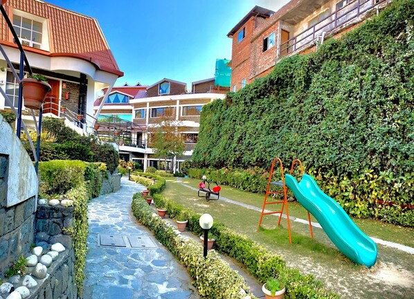 A courtyard with a children's play area, including a slide and swings, surrounded by neatly trimmed hedges and potted plants. - Marigold Sarovar Portico , Shimla. Heated pool & AC Rooms (Resort cottage and SPA) (Shimla)
