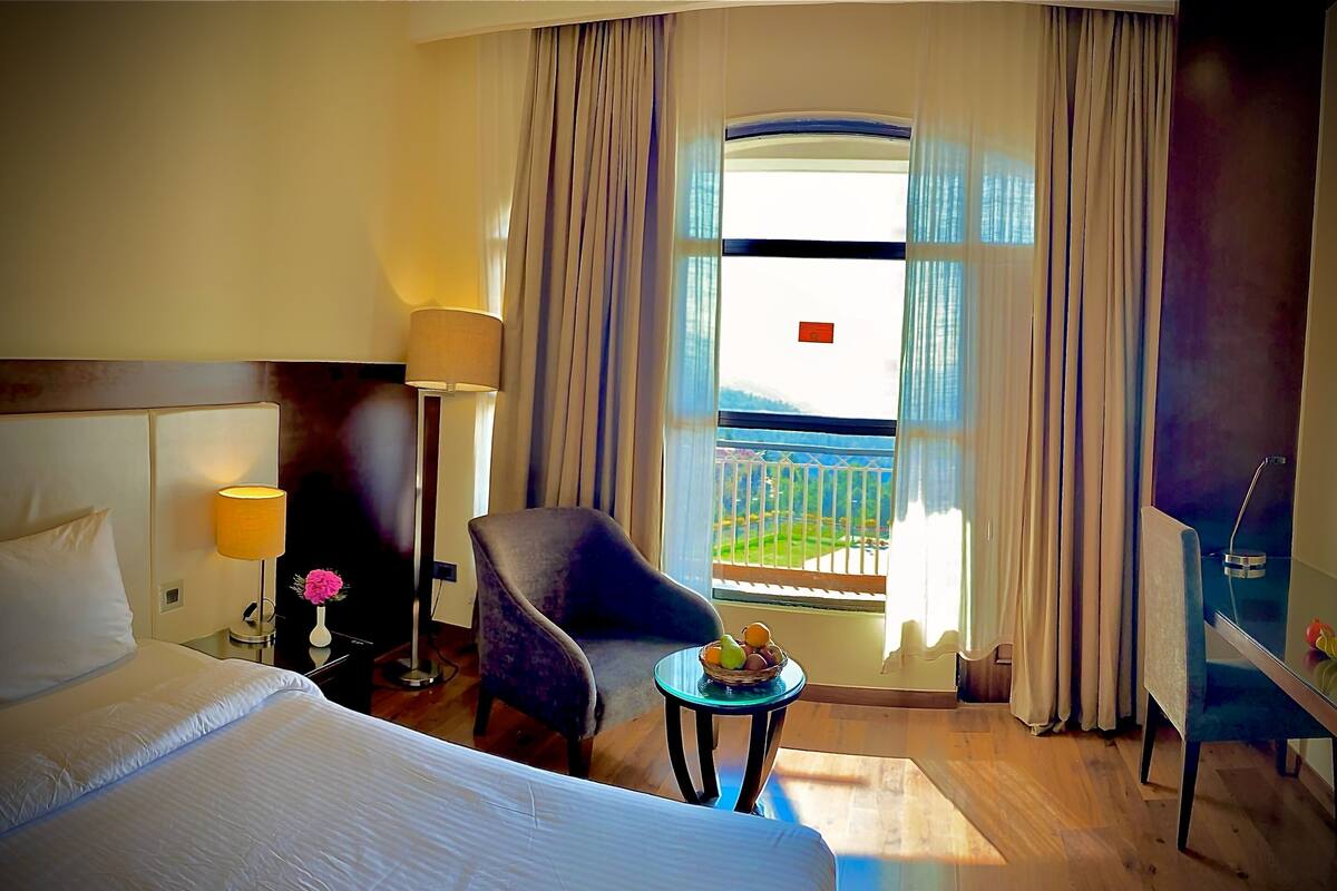 A hotel room with a bed, a chair, a small table with fruit, a lamp, and a window with curtains.