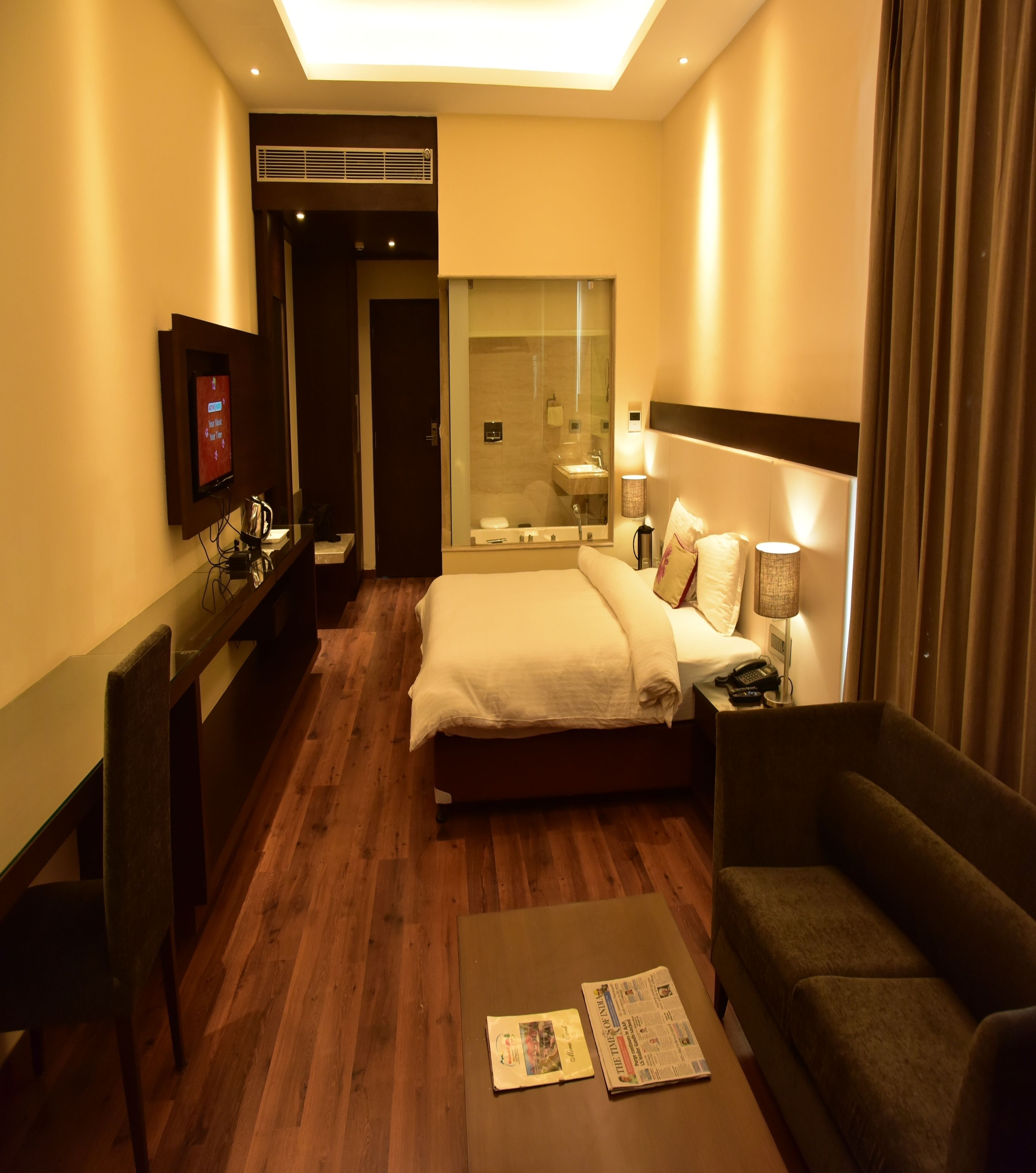 Habitación de hotel con cama, escritorio, sillón, televisión y baño visible a través de un panel de vidrio.