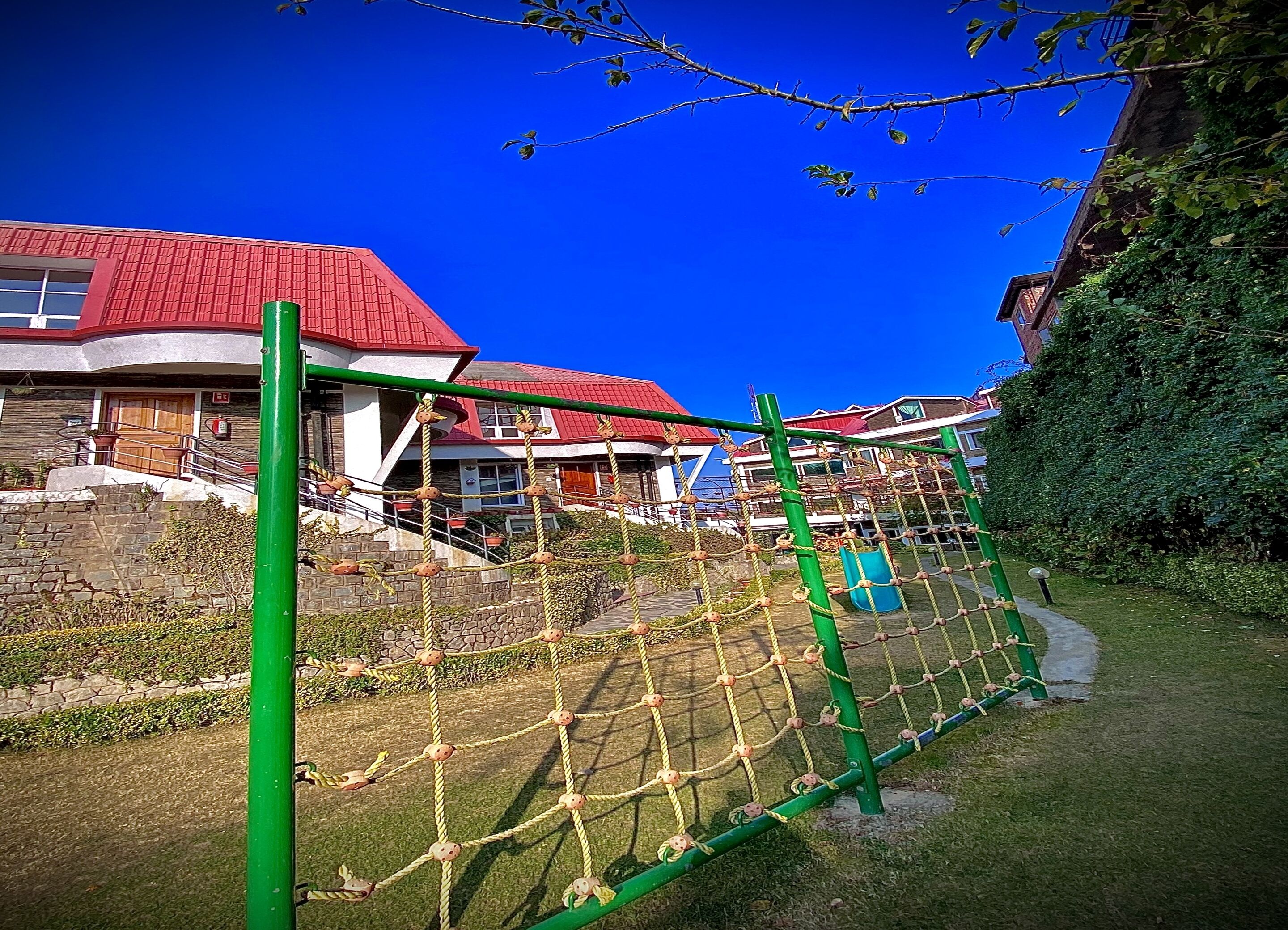 A playground with a rope net structure, a green pole, and a red-roofed building in the background.