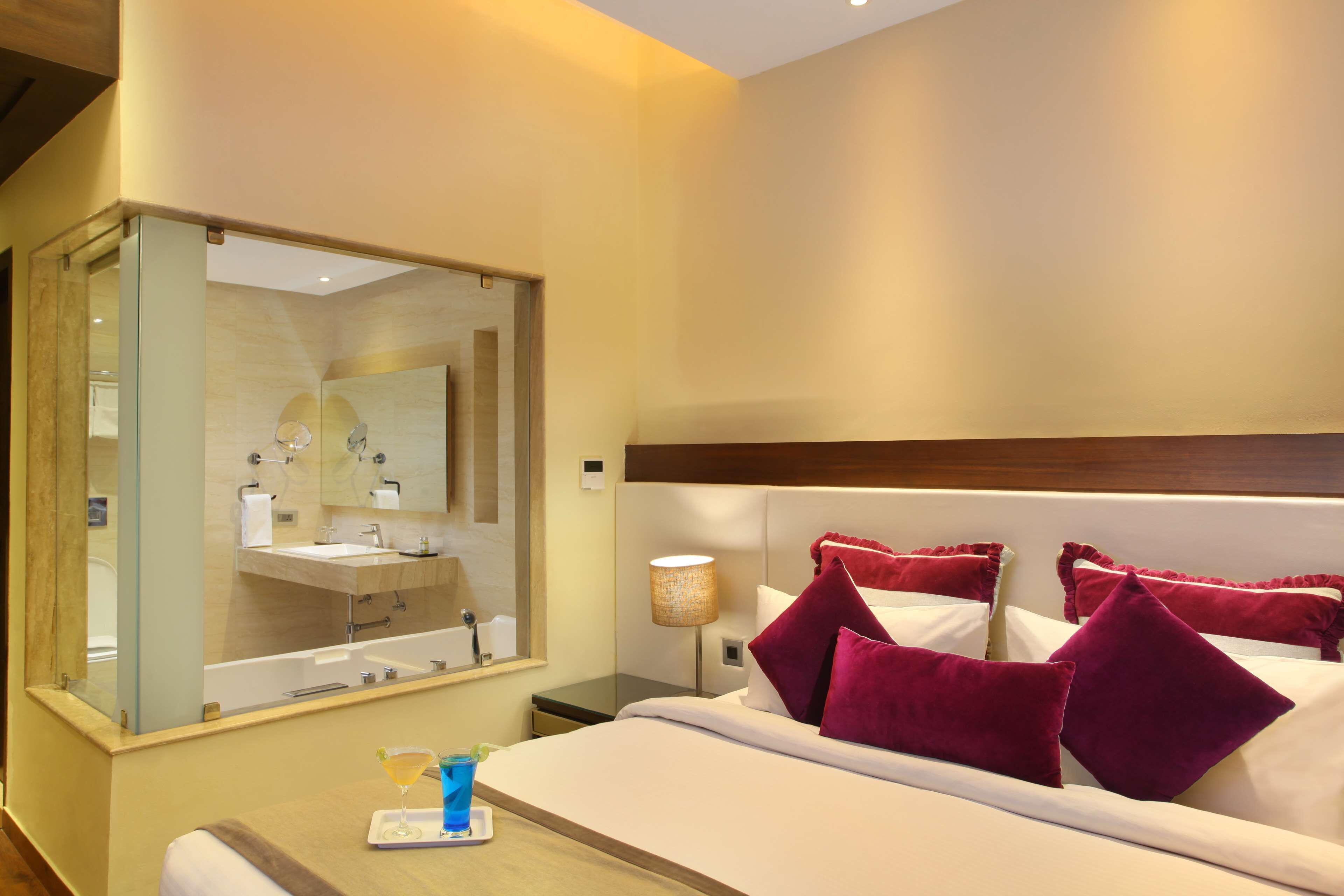 Habitación de hotel con un espejo grande, una cama con almohadas blancas y burdeos, una mesita de noche con lámpara y un baño visible a través del espejo.