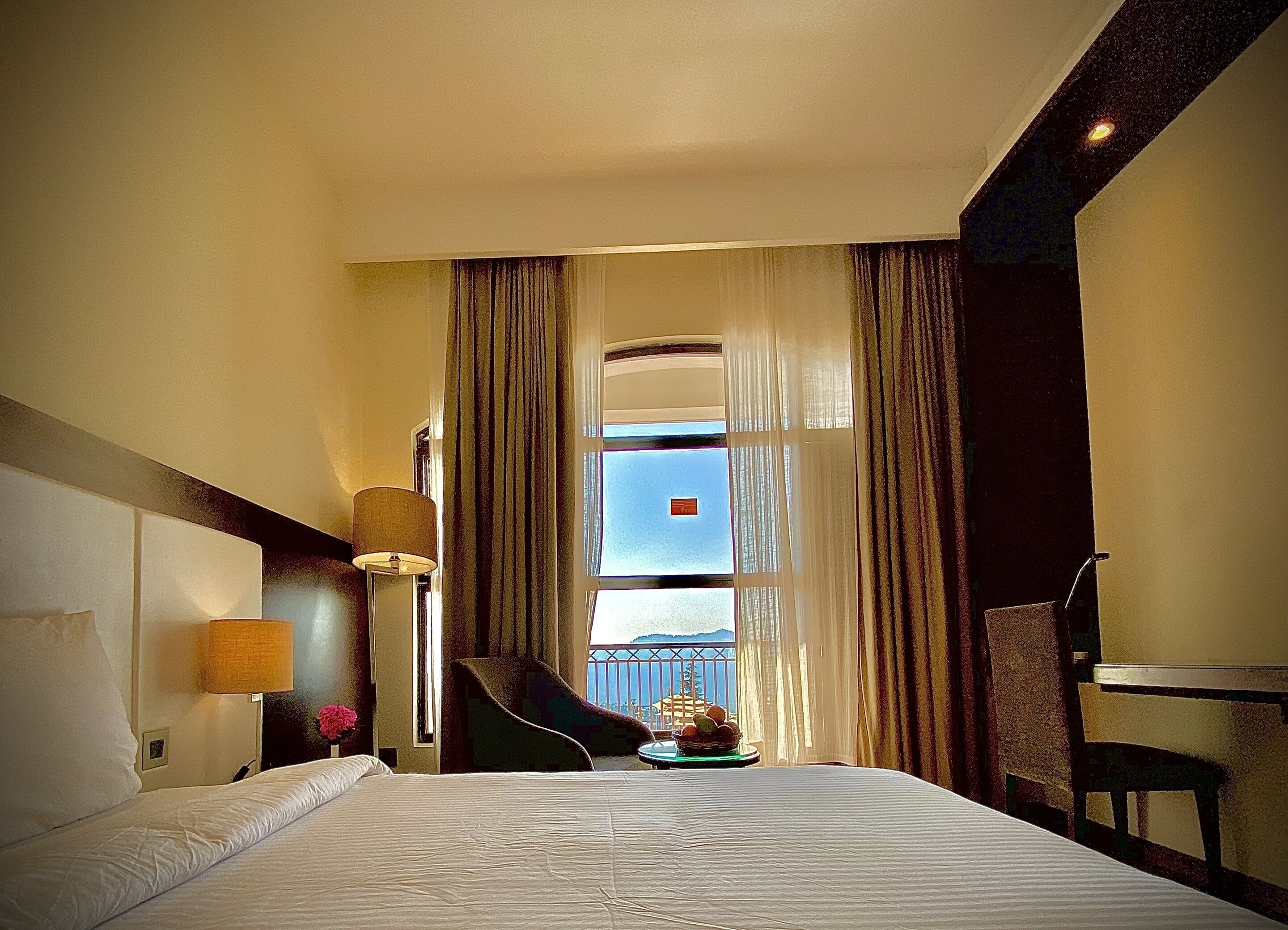 A hotel room with a large bed, a desk, a chair, a lamp, and a view of the outside.