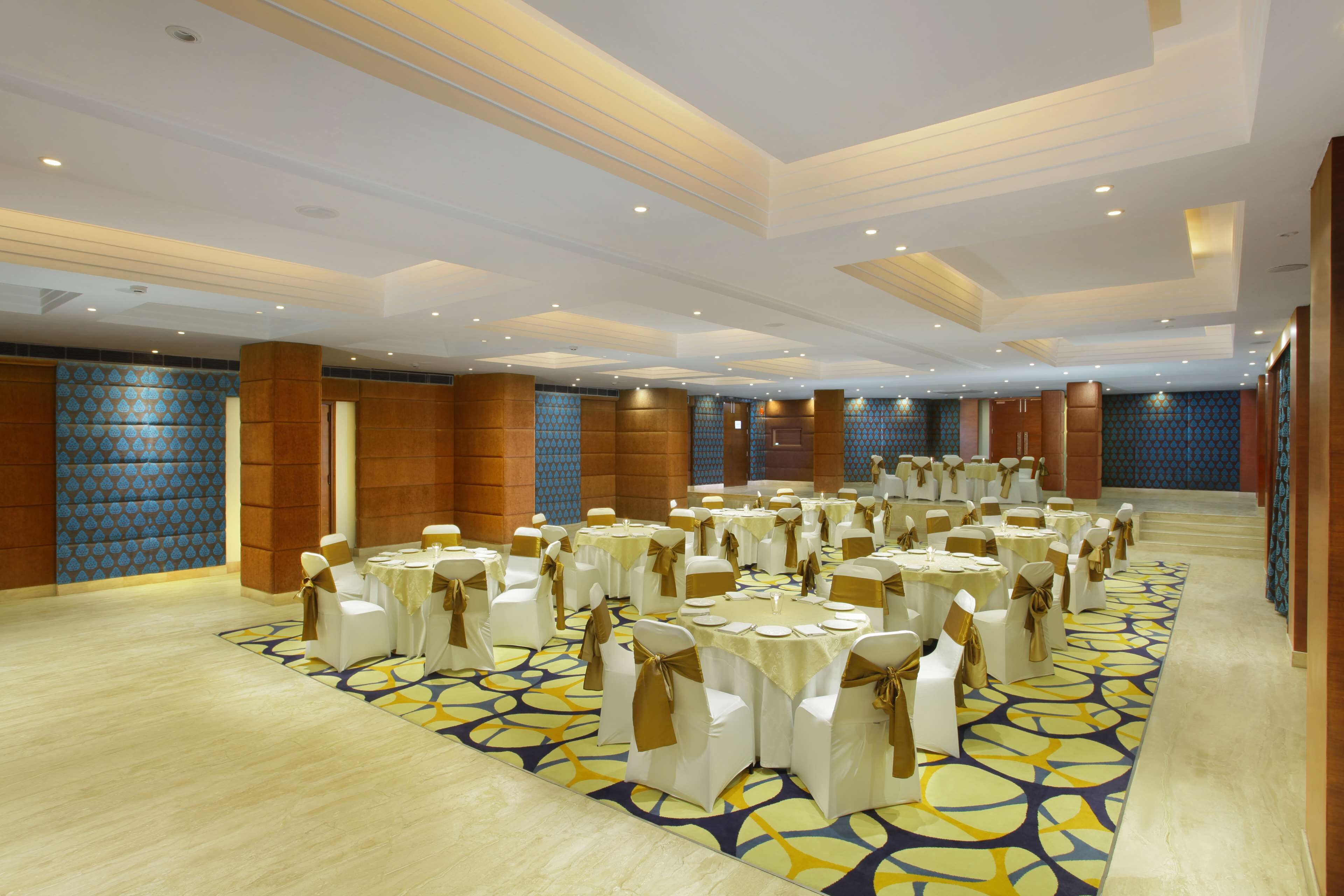 A large banquet hall with round tables and chairs, a patterned carpet, and a high ceiling with recessed lighting.