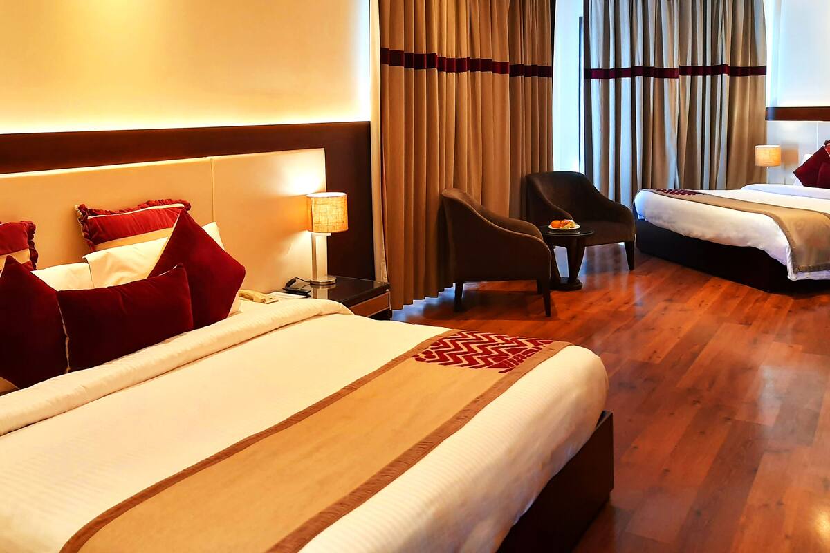 A hotel room with a large bed, a smaller bed, a desk with a lamp, a chair, and a view of the outdoors through curtains.