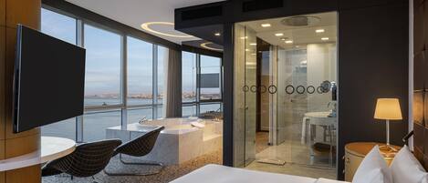 A modern hotel room with a large bed, a TV, a seating area with chairs, a bathroom with a sink and mirror, and a view of a cityscape through large windows.
